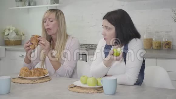 侧面看不高兴的高加索女人嫉妒的看着苗条的朋友吃美味的牛角面包和咀嚼苹果健康视频的预览图