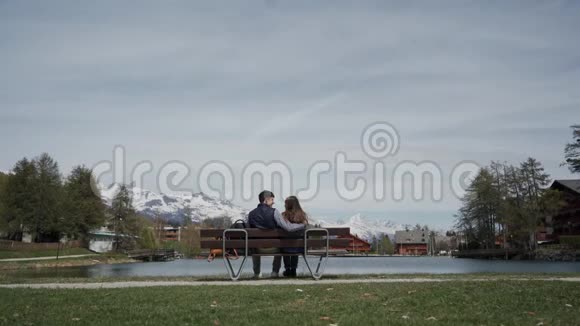后景浪漫的年轻夫妇坐在湖边的长凳上有着美丽的山景总计划视频的预览图