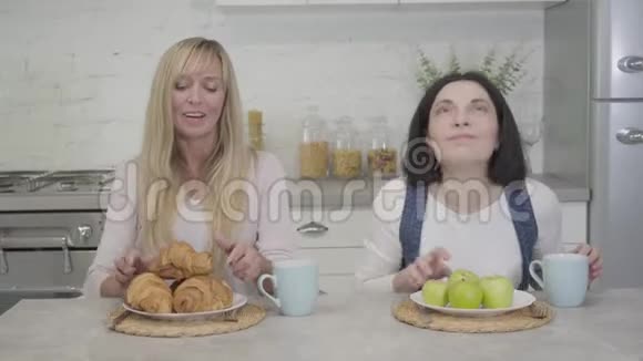 两个成年女性朋友闻和咬食物的肖像布鲁内特女士吃着绿苹果金发女郎享受着视频的预览图