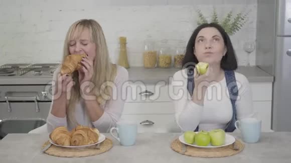 白种人女人吃健康苹果的肖像她的金发朋友喜欢吃不健康的羊角面包生活方式视频的预览图
