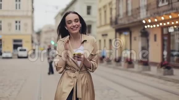 戴着耳机的漂亮女孩打开她的智能手机音乐和微笑站在城市背景视频的预览图