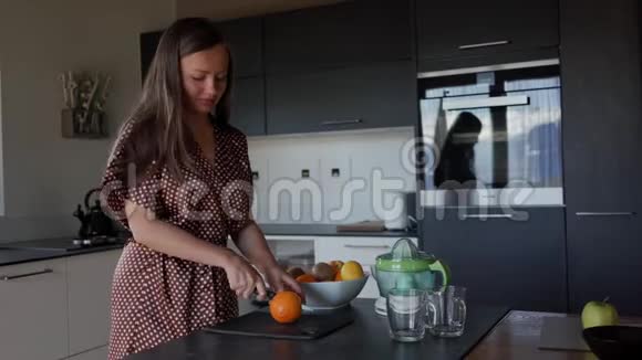 穿着棕色睡衣的微笑女人在厨房用刀子切橘子用来做新鲜果汁一般视频的预览图
