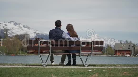 幸福的年轻夫妇坐在湖边的木凳上在瑞士阿尔卑斯山上有美丽的景色他抱着女朋友视频的预览图