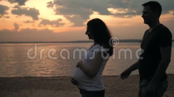 丈夫拥抱他深爱的怀孕的妻子站在日落的海滩上视频的预览图