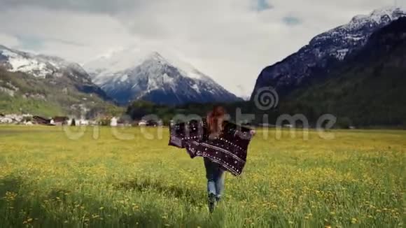 后视图风景如画的雪山上一位身穿花纹大衣的快乐女子张开双臂在蒲公英地上奔跑视频的预览图