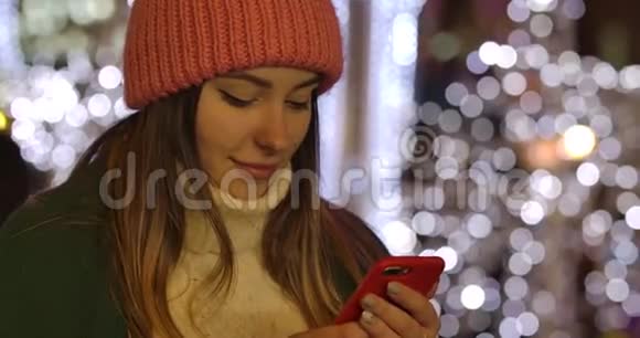 可爱的白种人女孩在冬天穿着智能手机和微笑的衣服年轻漂亮的女人花时间视频的预览图