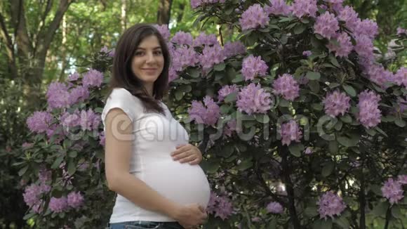 幸福的孕妇站在鲜花丛中用手抚摸她的胃视频的预览图
