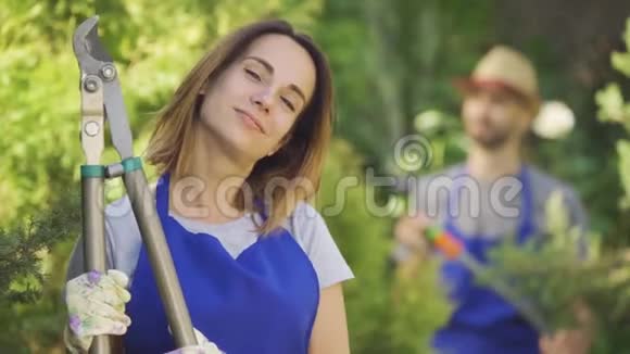 一幅美丽的白种人妇女的画像她伸出大拇指手里拿着大花园切割器在前景中而男人正在观望视频的预览图