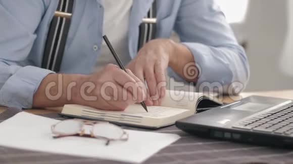 男性手拿笔在笔记本上做笔记在电脑上计算数字坐在桌旁视频的预览图