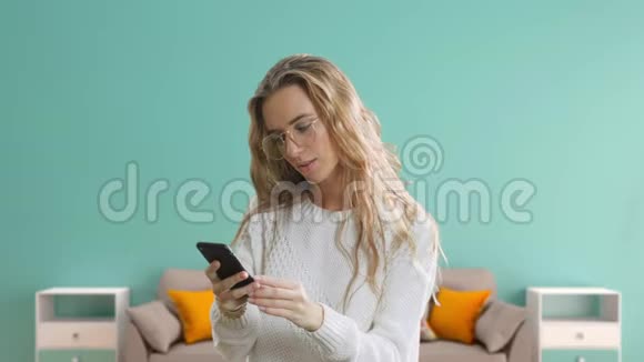 一个戴眼镜的快乐美丽女孩的肖像正在看一部手机在蓝色的房间背景下庆祝视频的预览图