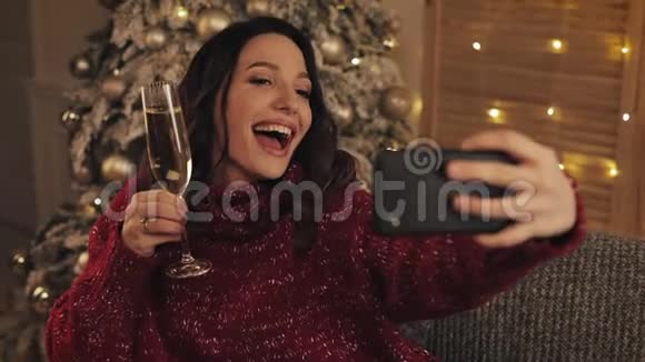 穿着冬季毛衣的年轻美丽的布鲁内特女士坐在家中的圣诞树旁背景是拿着香槟视频的预览图