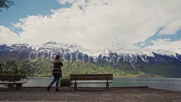 木制长凳可观赏山景和蓝色湖泊穿着温暖图案外套的可爱女孩来到长凳上坐在上面视频的预览图