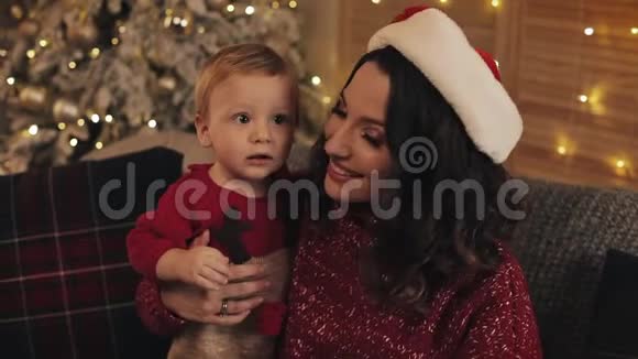 靠近微笑美丽的妈妈戴着圣诞帽抱着她的小男孩坐在客厅附近的沙发上视频的预览图