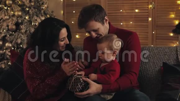 爸爸妈妈和小儿子坐在圣诞树附近的沙发上妈妈拍手爸爸拿着礼物盒宝贝视频的预览图