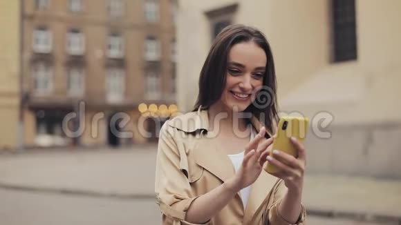 穿着米色壕沟的棕色头发的漂亮年轻女孩在智能手机上对着老城区微笑视频的预览图
