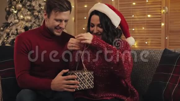 可爱的年轻夫妇一起完成圣诞礼物乐队磁带和绑在碗坐在沙发附近视频的预览图