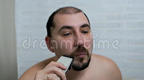一个男人在家里用电动剃须刀刮胡子和胡子在镜子前脱毛男人照顾视频的预览图