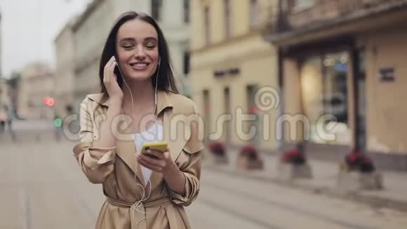 戴着耳机的漂亮女孩喜欢听音乐手持智能手机微笑着在城市散步视频的预览图