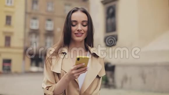 穿着米色壕沟的漂亮年轻女人用她的智能手机和微笑迫使她在旧城扫头发视频的预览图