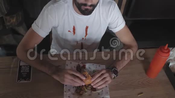 一个留着胡子的人穿着一件白色T恤上面涂着番茄酱坐在一张木桌旁拿着一个汉堡包男人吃汉堡包视频的预览图