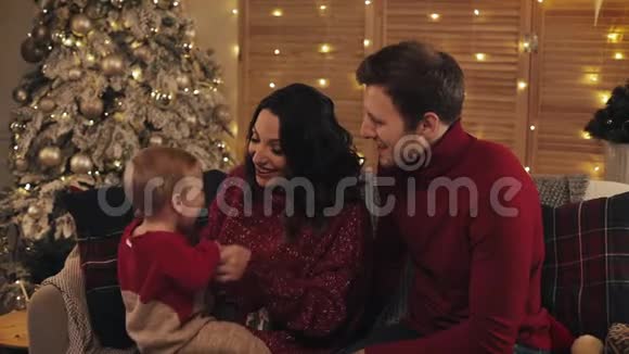 快乐的父母在舒适的家背景下坐在圣诞树旁的沙发上抱着他们兴奋的宝贝儿子玩视频的预览图