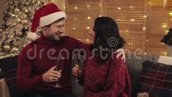 幸福的年轻夫妇坐在沙发上靠近圣诞树在舒适的家背景下闪烁着香槟酒的玻璃视频的预览图