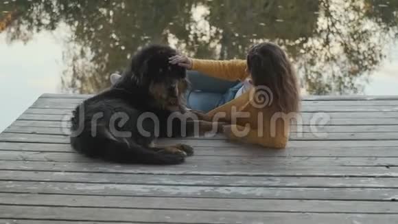 一个穿黄色夹克的年轻女人躺在码头上抚摸着一只大狗水中树木的倒影视频的预览图