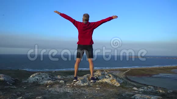一个穿着红色夹克衫的瘦长男子迎风挺立在高山上描绘了一个张开双臂的飞行视频的预览图