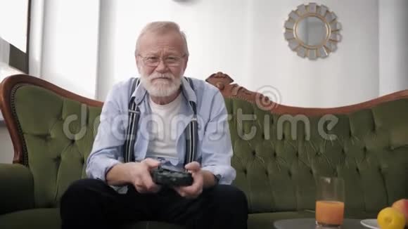 现代的有胡子和视力眼镜的祖父玩家坐在沙发上玩电子游戏拿着操纵杆视频的预览图