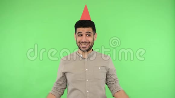 有魅力的积极男人头上戴着派对锥衬衫上戴着滑稽的帽子室内摄影棚拍摄绿色背景视频的预览图