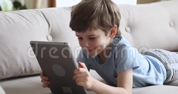 快乐可爱的学龄前男孩独自躺在沙发上用平板电脑视频的预览图