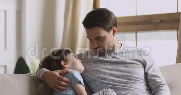 爱心爸爸抱着学龄前儿子在沙发上进行信任对话视频的预览图