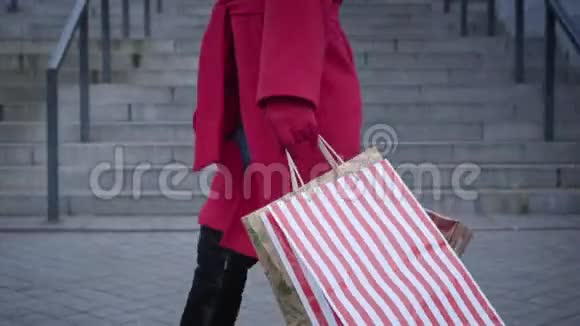 在城市里穿着红色外套和手套带着购物袋走路的无法辨认的女人的侧面景色年轻时尚女士视频的预览图