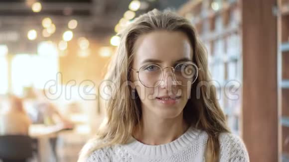 在大学图书馆背景下戴眼镜的聪明女孩看相机的肖像一个聪明的少年微笑着视频的预览图