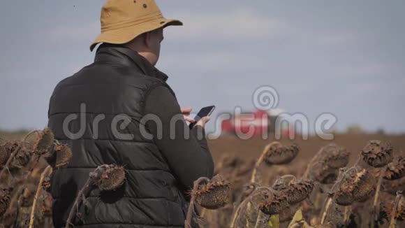 英俊的农民与智能手机站在田间向日葵与联合收割机的背景现代概念视频的预览图