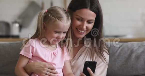 快乐妈妈抱着小女儿坐在沙发上用电话视频的预览图