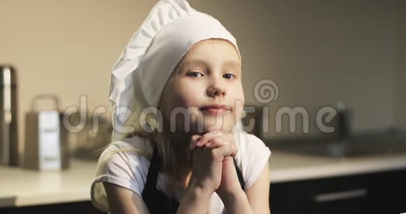 可爱的女孩穿着围裙和一顶白色的帽子站在厨房里做饭视频的预览图