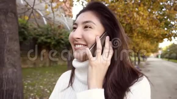 慢动作亚洲快乐女孩穿着白色毛衣用智能手机说话在公园里散步视频的预览图