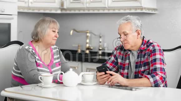 在厨房吃早餐时一对老夫妇微笑着用智能手机说话视频的预览图