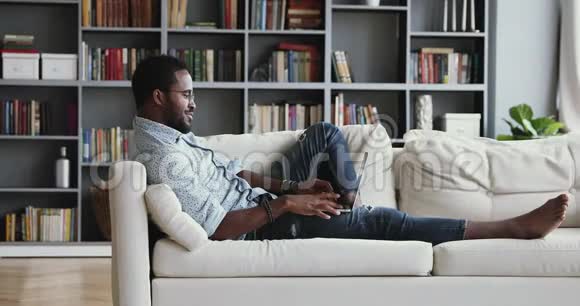 千禧一代非洲男人坐在沙发上用笔记本电脑视频的预览图