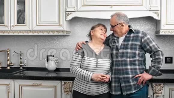 微笑着美丽的老人和男人拥抱微笑着摆姿势在厨房看着镜头视频的预览图