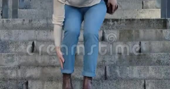 女性白种人的腿部特写因为脚踝疼痛而走下楼梯并停止穿牛仔裤和棕色衣服的女人视频的预览图
