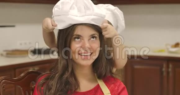 一个成年白种人女人的特写镜头看着镜头微笑小女孩把厨师的帽子戴在妈妈的头上视频的预览图
