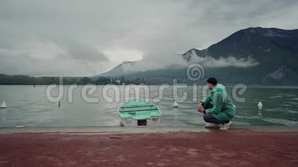 穿着薄荷雨衣的人坐在山上的一个湖上靠近一艘颜色相同的停泊船雨天安纳西湖视频的预览图