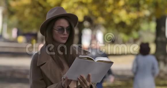 年轻的高加索女学生穿着优雅的棕色衣服和太阳镜在秋天公园看书聪明的人视频的预览图