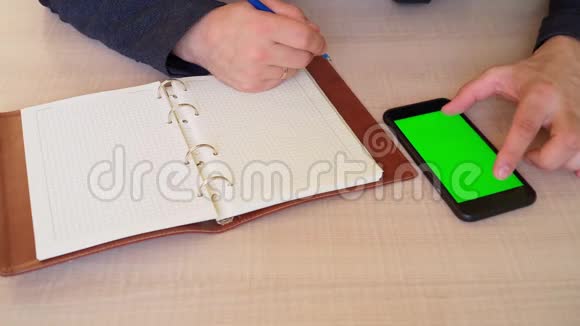 特写镜头那家伙在笔记本上记笔记手里拿着智能手机绿色屏幕男人拿着手机的色度键视频的预览图