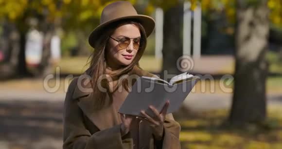 年轻聪明的高加索女人穿着优雅的棕色衣服和太阳镜在秋天公园看书魅力视频的预览图