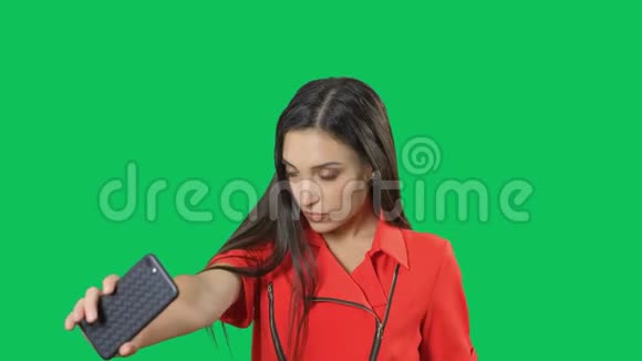 穿着红色连衣裙的快乐年轻女子正在绿色屏幕背景上拍摄自拍照片移动摄影摄像应用软件视频的预览图