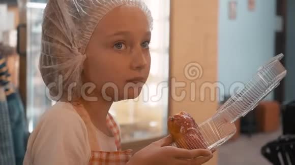 穿着一次性帽子和厨房围裙吃甜甜圈的少女厨师女孩在糕点店吃新鲜甜甜圈肖像视频的预览图