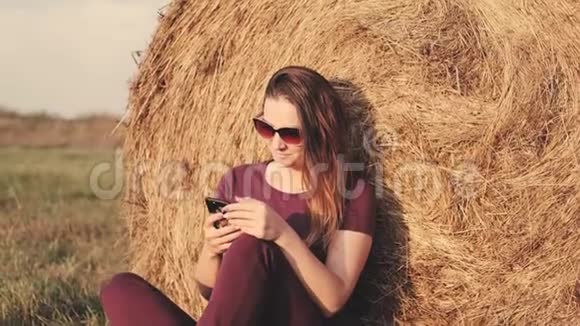 一个拿着智能手机的女人坐在干草堆旁边的田野里视频的预览图